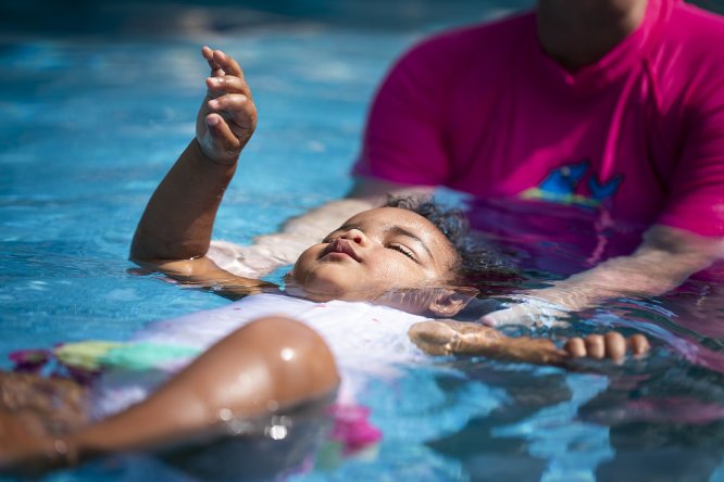 Young child floating on their back in a swimming pool during an ISR-style lesson, supported by an instructor in a bright pink swim shirt.